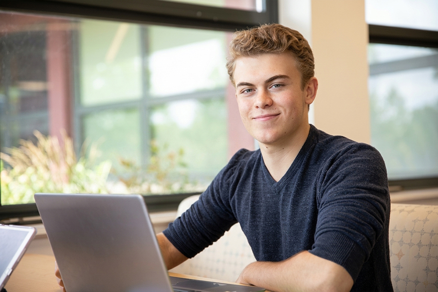 Student sitting at a laptop by a window, smiling at the camera