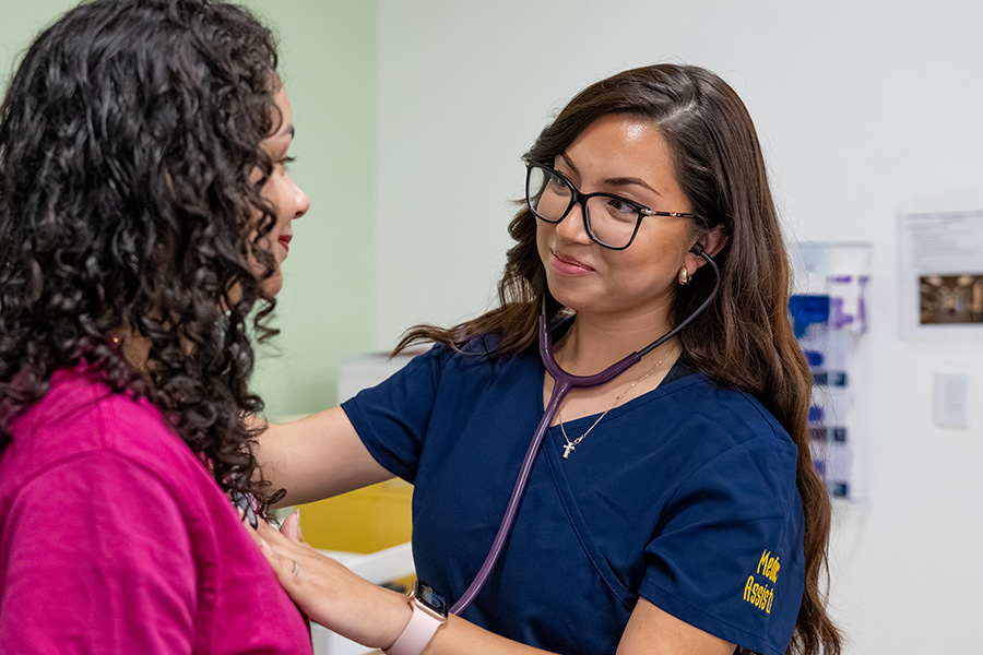 Medical assistant student in navy scrubs using a stethoscope to listen to a patient's heart in an exam room